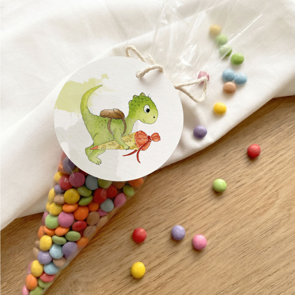 Runder Anhänger Einschulung mit Dino Aquarell Motiv an Spitztüte voller bunter Schokolinsen auf hellem Holztisch.