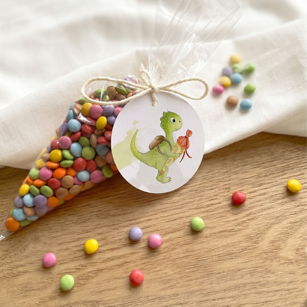 Kleiner Anhänger Einschulung mit Dino Aquarell Motiv befestigt an Klarsichtbeutel mit bunter Linsenfüllung.