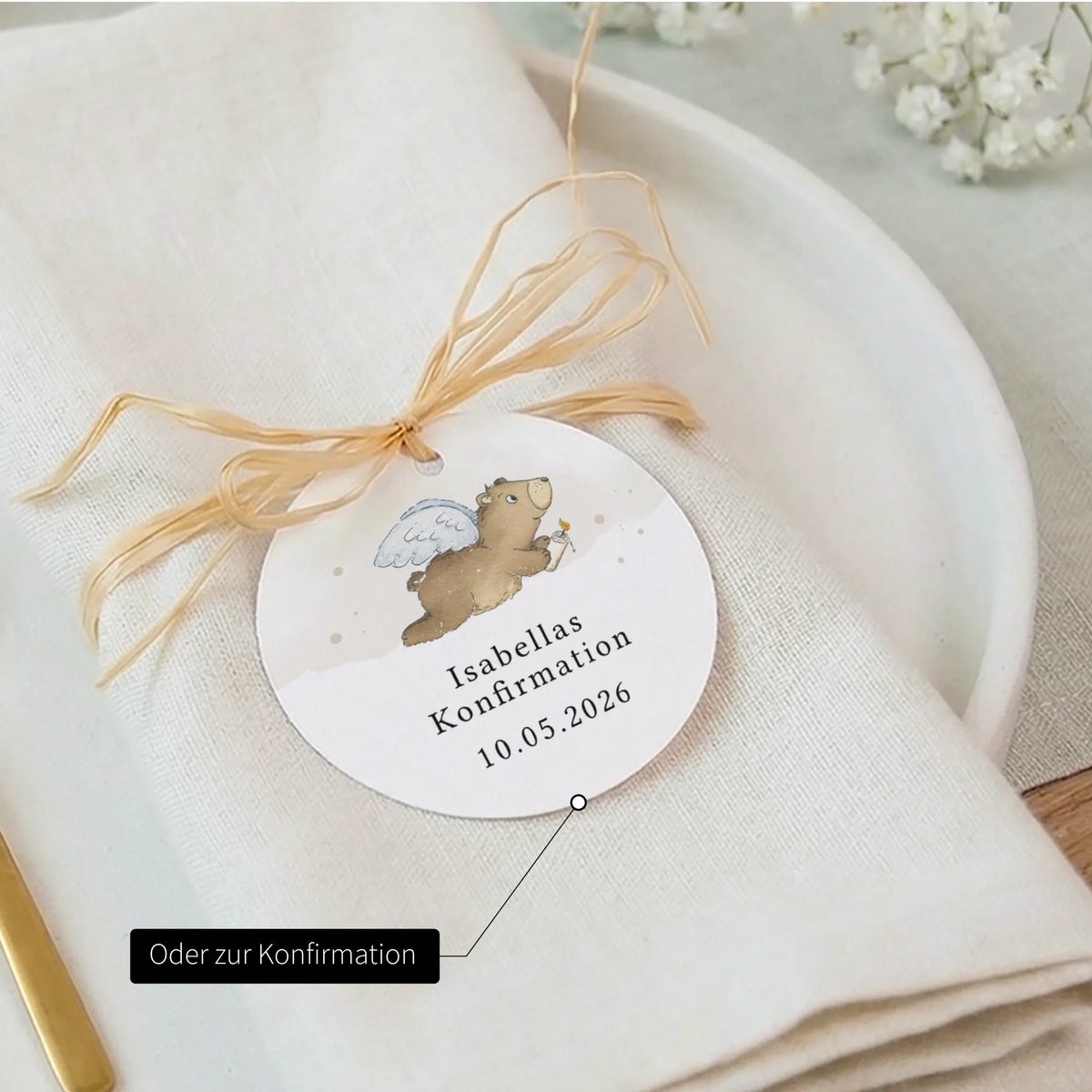 Personalisierter runder Anhänger zur Konfirmation mit Aquarell Motiv Engelbär, Aufschrift Isabellas Konfirmation 10.05.2026, auf gefalteter Serviette.
