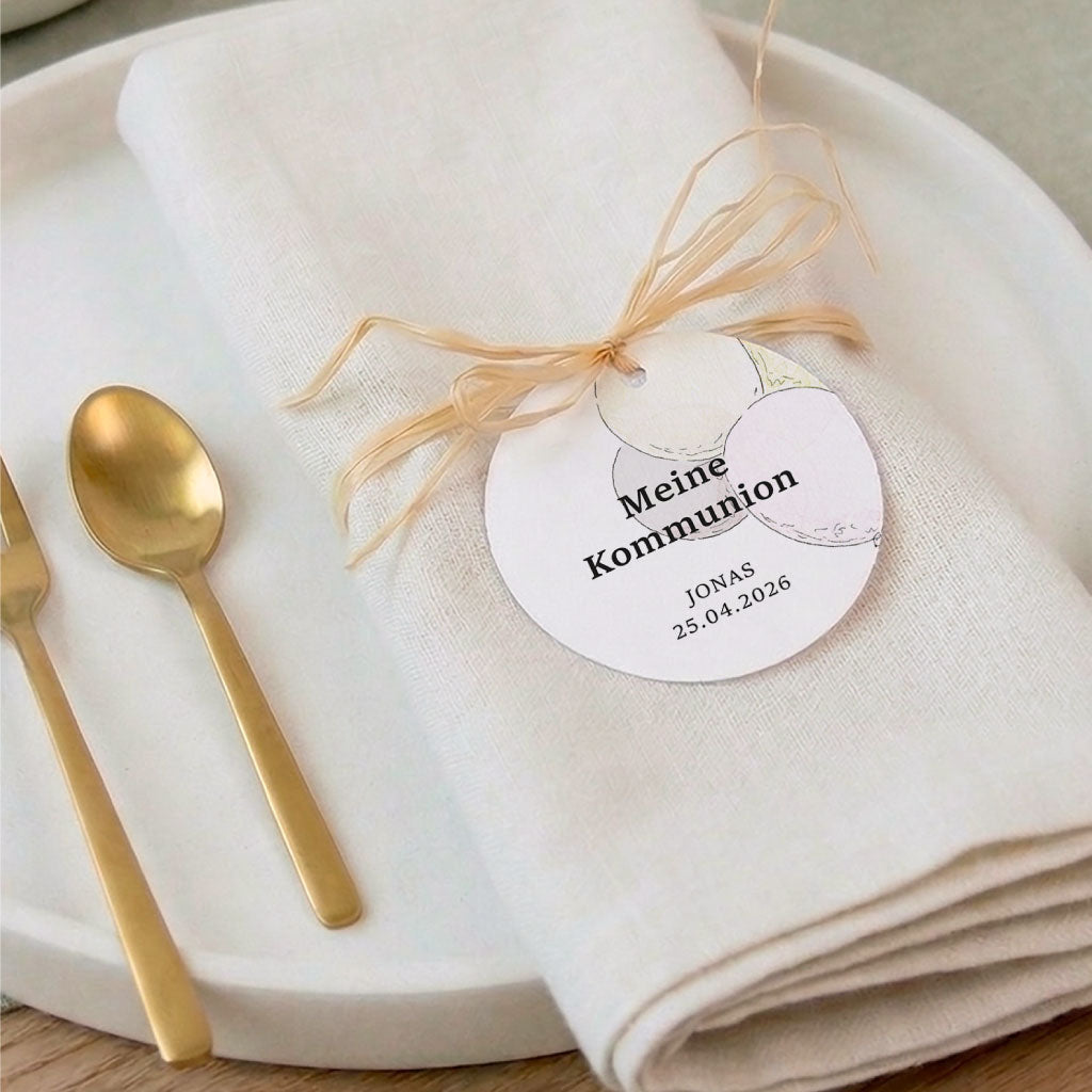 Runder Anhänger mit Luftballon Aquarell Motiv als Dekoration auf einer Serviette mit goldenem Besteck.