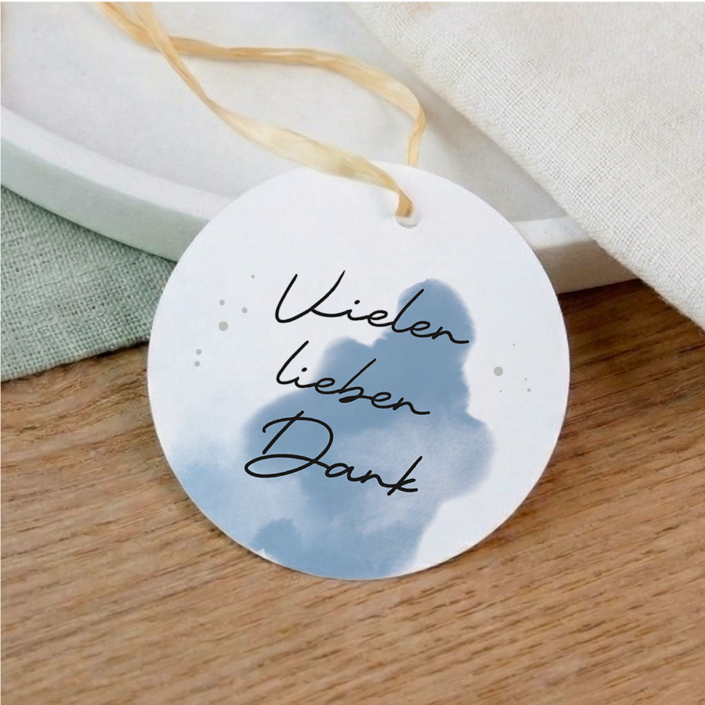 Runder Anhänger für Taufe und Kommunion mit Aufschrift Vielen lieben Dank und blauem Aquarell Motiv auf einem Holztisch.