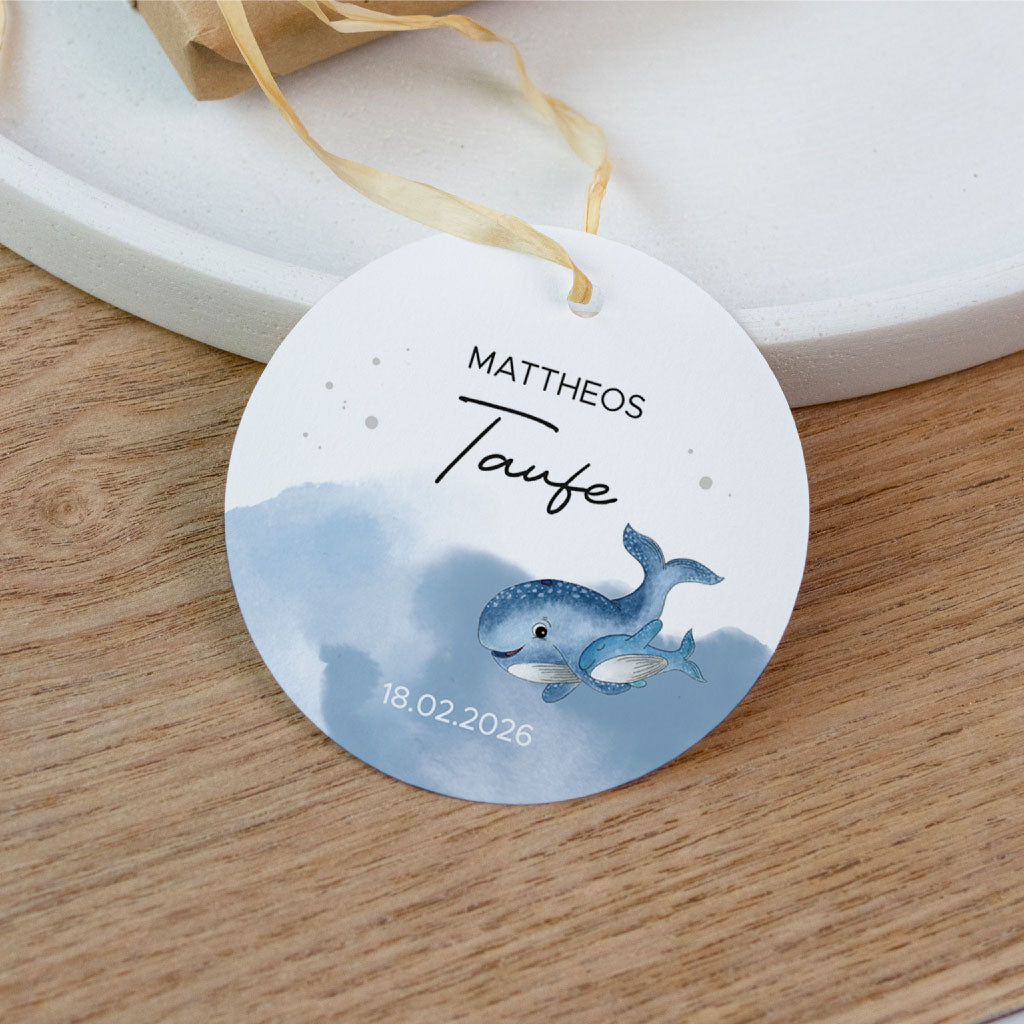 Anhänger zur Taufe mit Name und Datum liegend auf einem Holztisch neben einem kleinen Geschenk.
