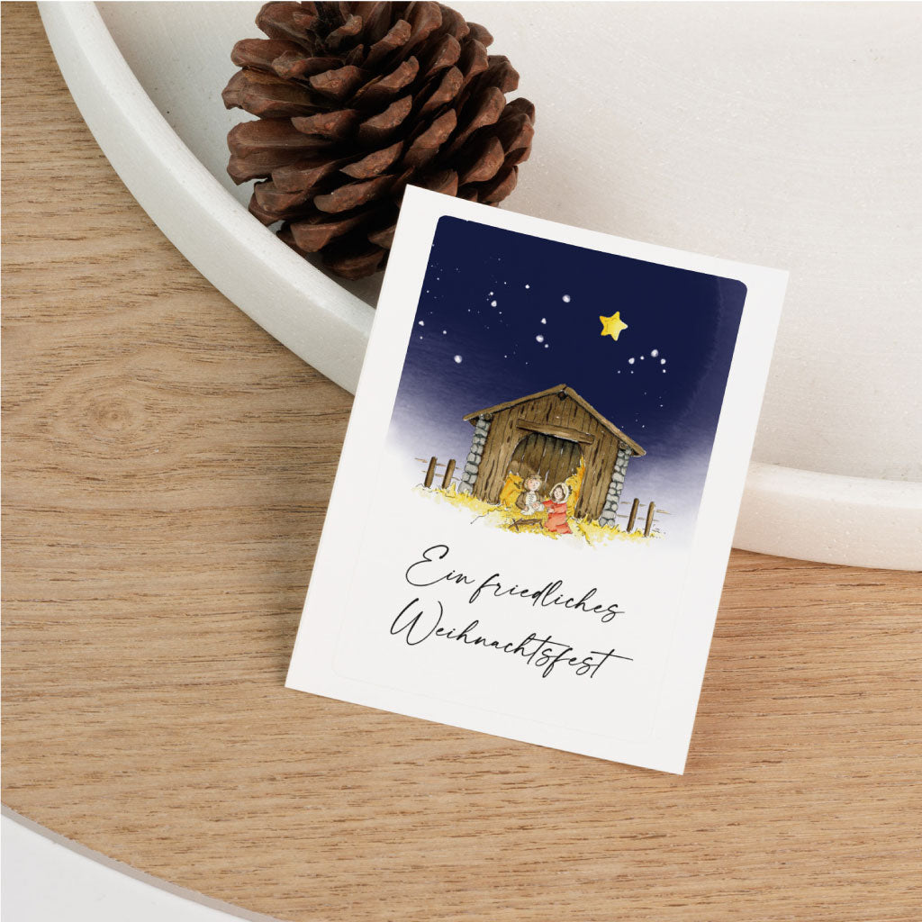 Sticker mit Krippenmotiv auf hellem Holzuntergrund, liebevoll illustriert in Aquarell, ideal für Grabkerzen oder als weihnachtlicher Aufkleber mit christlicher Botschaft.