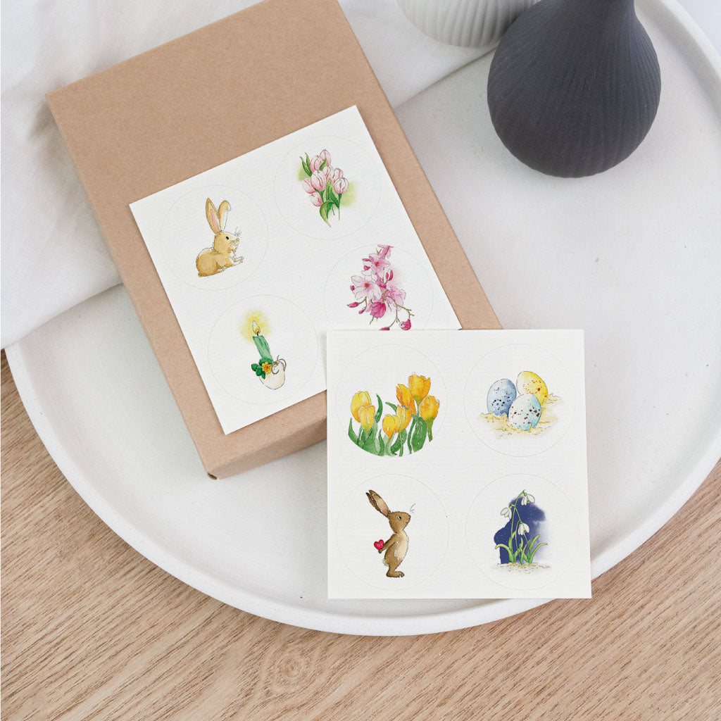 Handgemaltes Aufkleber Set Ostern mit runden Stickern: Osterhase, Ostereier und Frühlingsblumen als Aquarelle auf Karten, dekorativ auf Geschenkbox arrangiert.