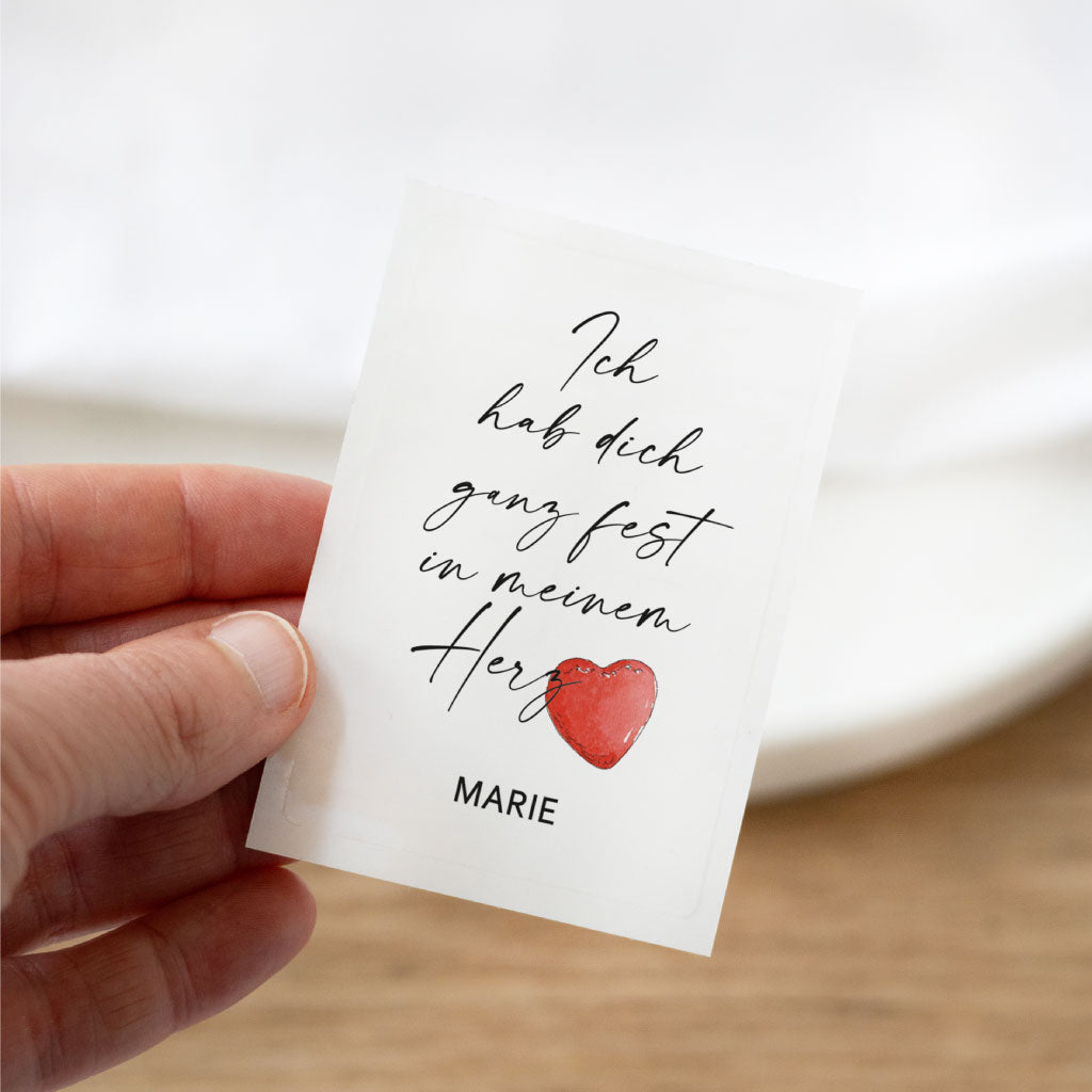 Transparenter Grabkerzen-Aufkleber mit rotem Aquarell-Herz und Name „Marie“ – detailreiche Nahaufnahme in der Hand, ideal als Geschenkidee zum Valentinstag.
