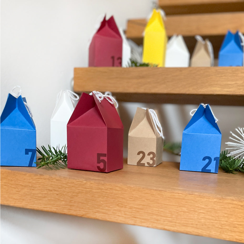 Bastel-Adventskalender aus kleinen Häuschen in verschiedenen Farben auf einer Holztreppe dekoriert – kreatives DIY-Projekt für die Adventszeit.