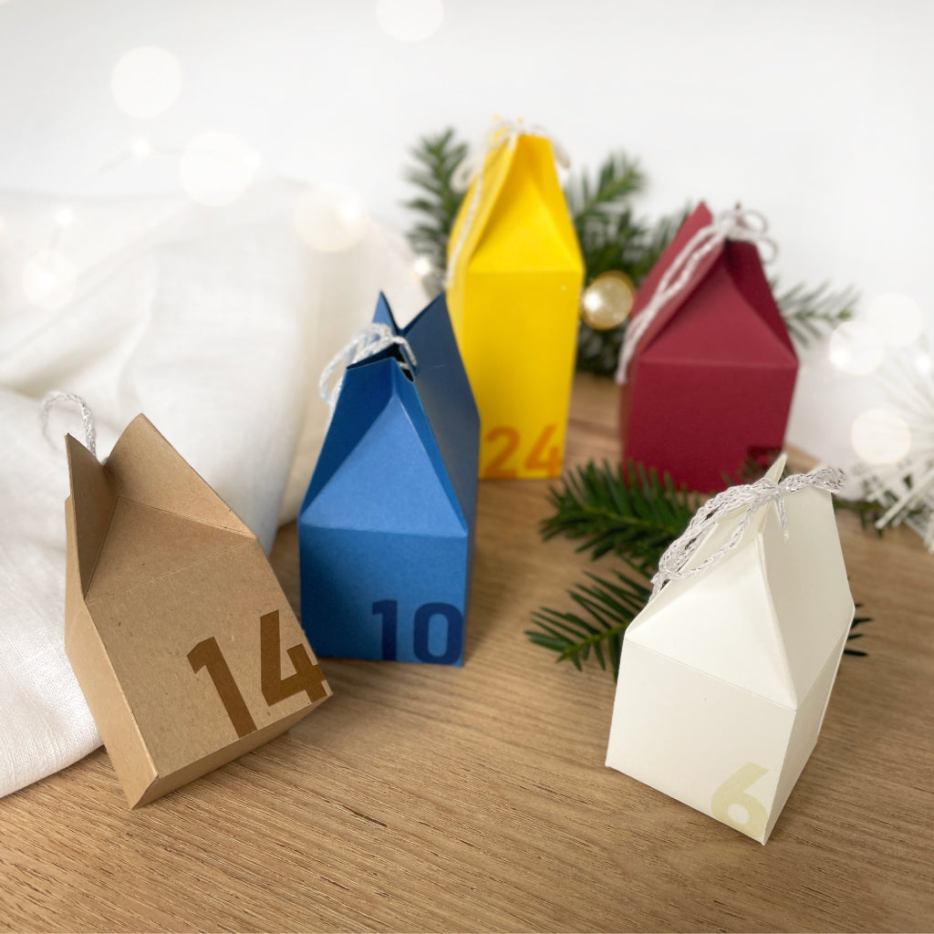Fünf bunte Adventshäuschen aus Karton als Teil eines DIY Sets, liebevoll arrangiert mit Tannenzweigen – perfekt für kreative Bastelfans.