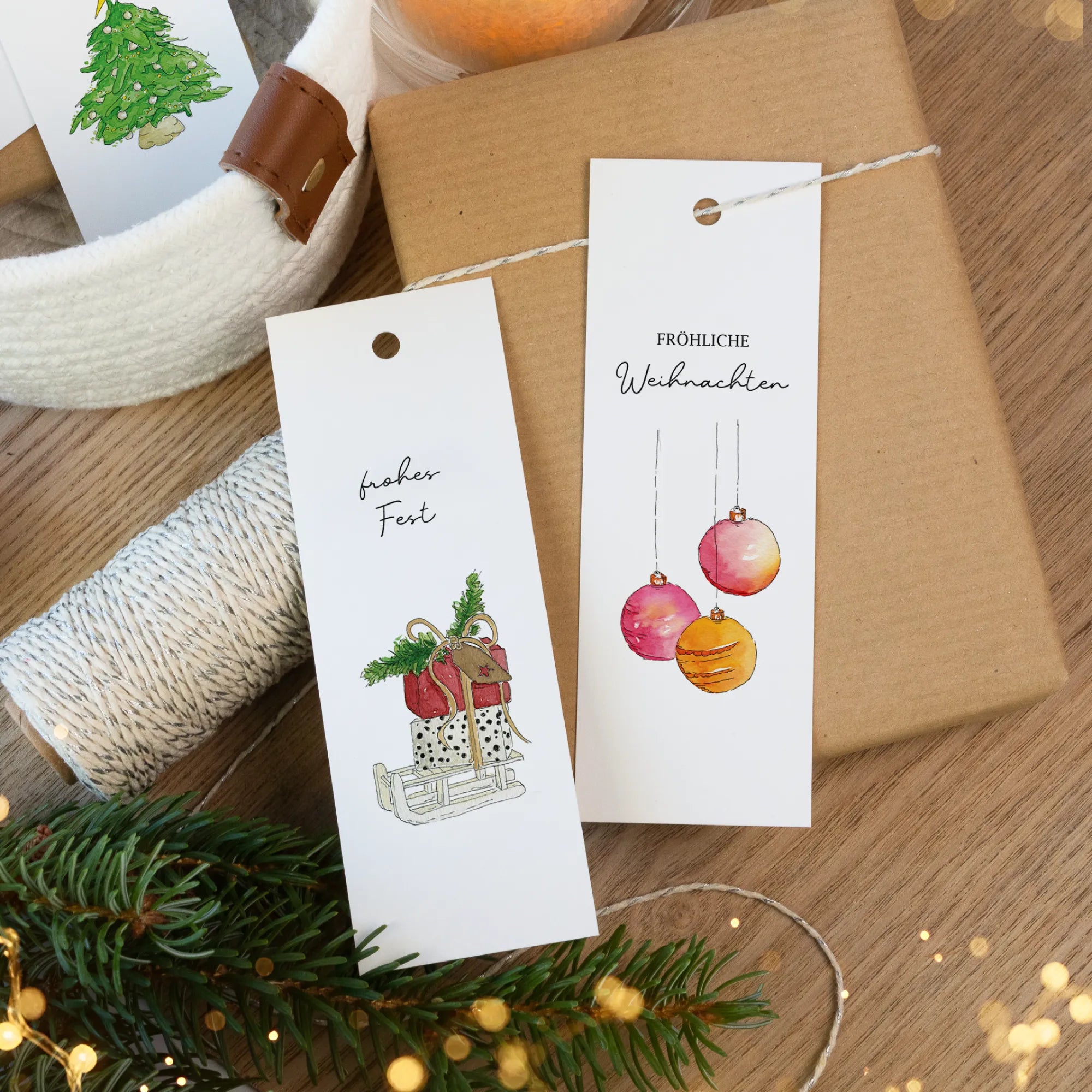 Verschiedene Geschenkanhänger mit Aquarell Motiv von Schlitten und Christbaumkugeln neben einer Tannenzweig Dekoration auf Holz.