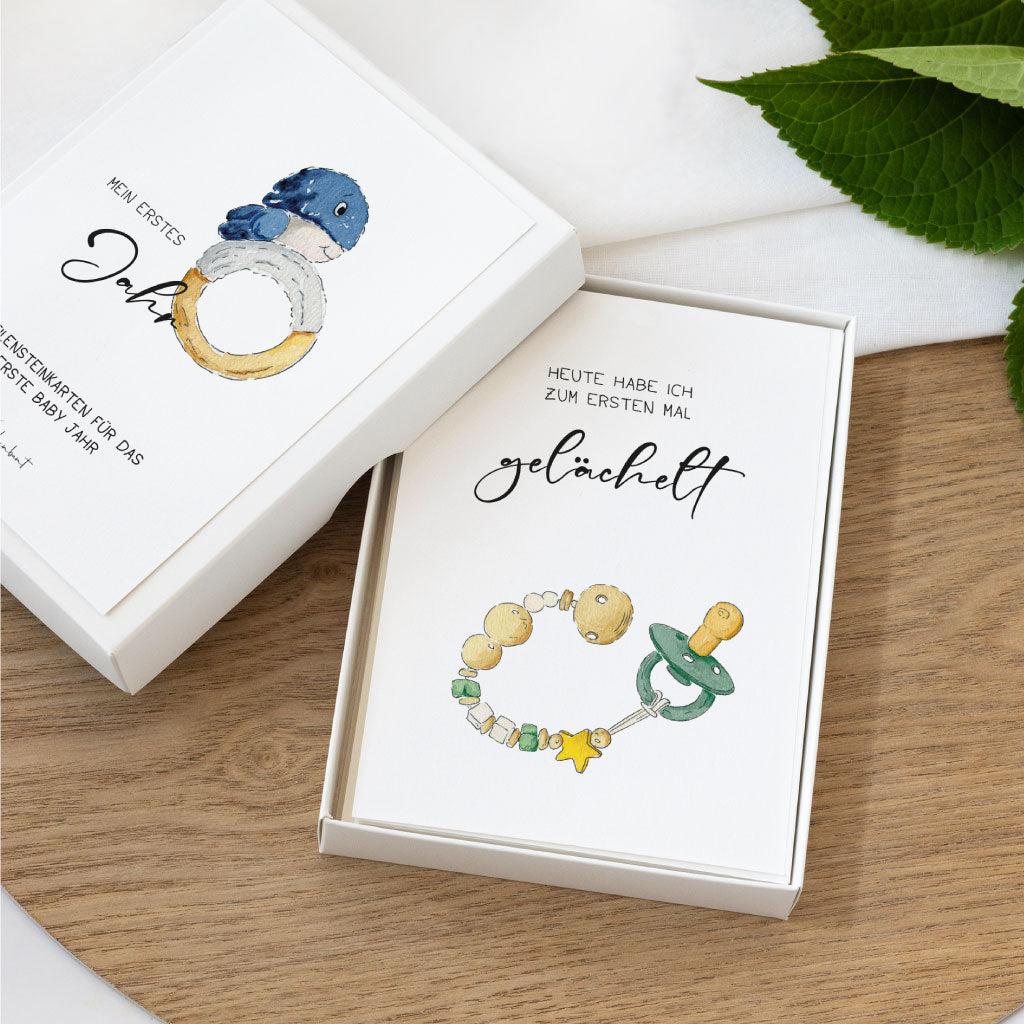 Zwei Meilensteinkarten im Geschenkkarton mit Aquarellzeichnungen: eine mit Greifring und Wal für „Mein erstes Jahr“, eine mit Schnuller für „Zum ersten Mal gelächelt“, als digitaler Sofort-Download.
