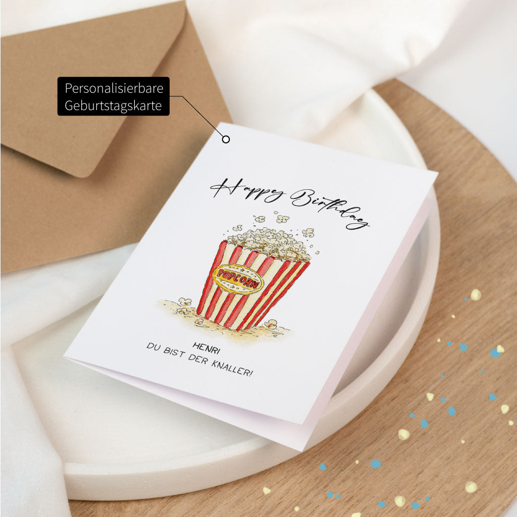 Personalisierbare Geburtstagskarte mit Aquarellillustration einer Popcorntüte und handschriftlichem „Happy Birthday“-Schriftzug, daneben ein Kraftpapier-Umschlag.