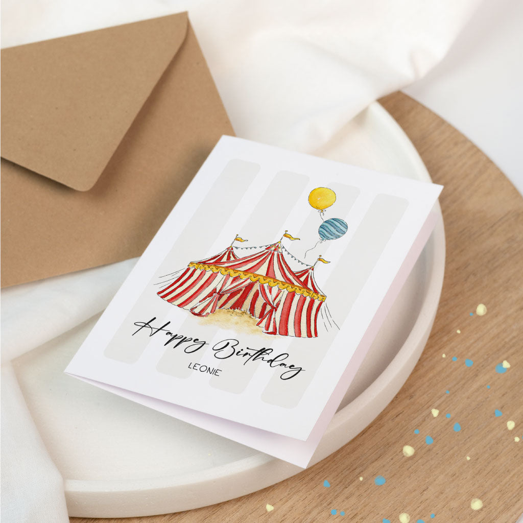 Geburtstagskarte im Zirkusdesign mit Aquarell-Illustration und Luftballons, personalisiert mit dem Namen „Leonie“, auf hellem Untergrund mit Umschlag arrangiert.