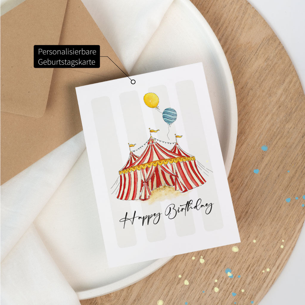 Personalisierbare Geburtstagskarte mit handgemaltem Zirkuszelt in Aquarelloptik und „Happy Birthday“-Schriftzug, auf einem Teller mit Streudeko platziert.
