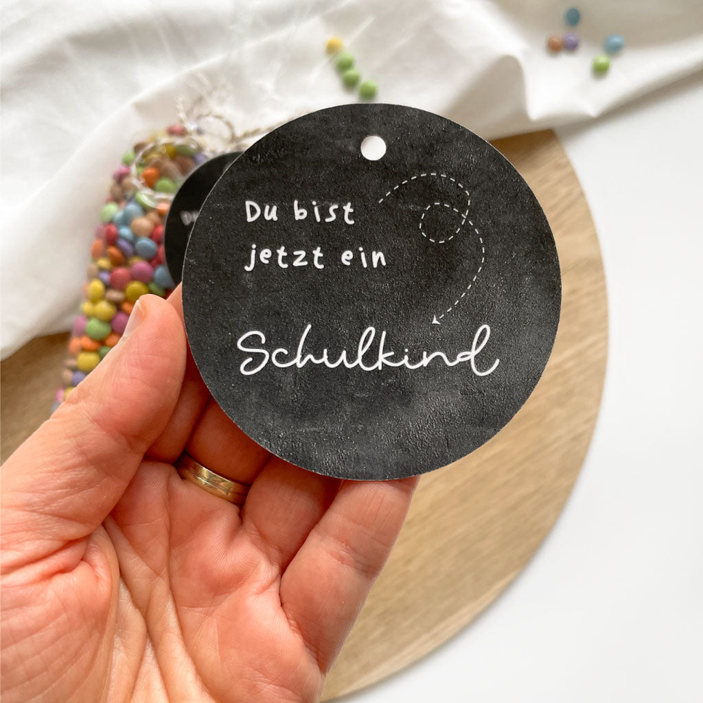 Anhänger wird in Hand gehalten, im Hintergrund Spitztüten mit bunten Schokolinsen