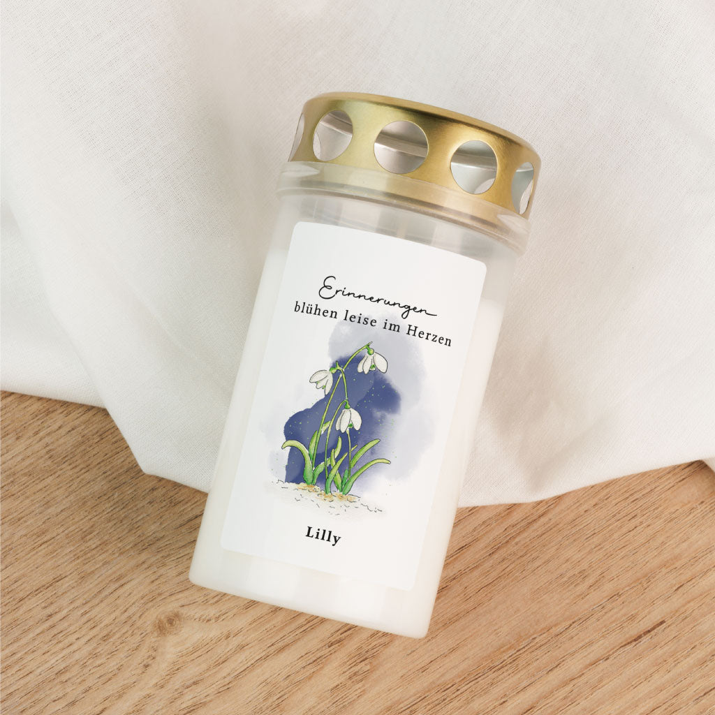 Grabkerze mit goldfarbenem Deckel und Aquarell-Schneeglöckchen, auf hellem Holz und Stoff dekorativ arrangiert – personalisierbar mit Namen.