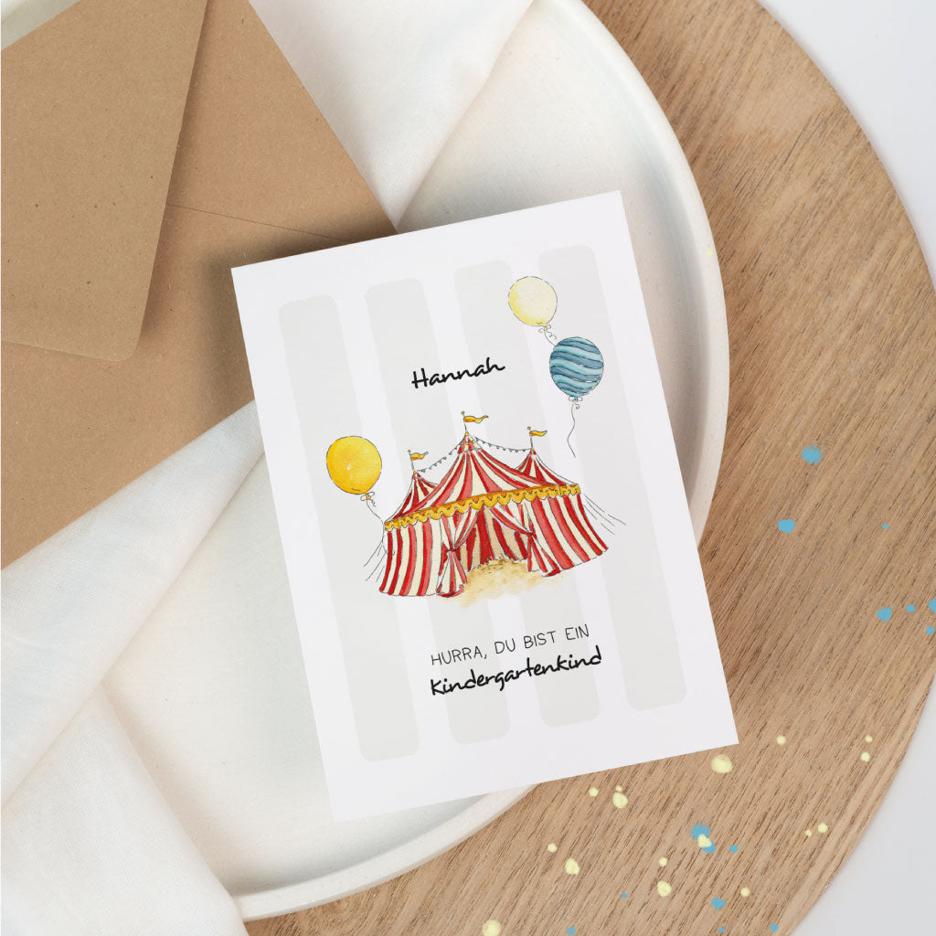 Personalisierbare Glückwunschkarte zum Kindergarten-Beginn mit Zirkusmotiv: rotes Zirkuszelt und Ballons, Name „Hannah“, Text „Hurra, du bist ein Kindergartenkind“