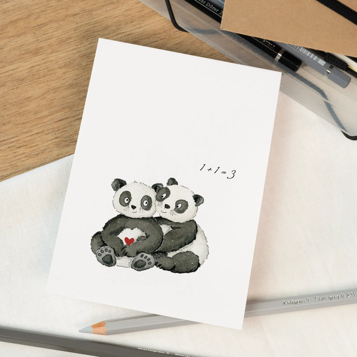 Schwangerschaftskarte mit handgemalten Pandas die sich umarmen und rotem Herz, Aufschrift 1+1=3, liegt auf Zeichenmaterialien