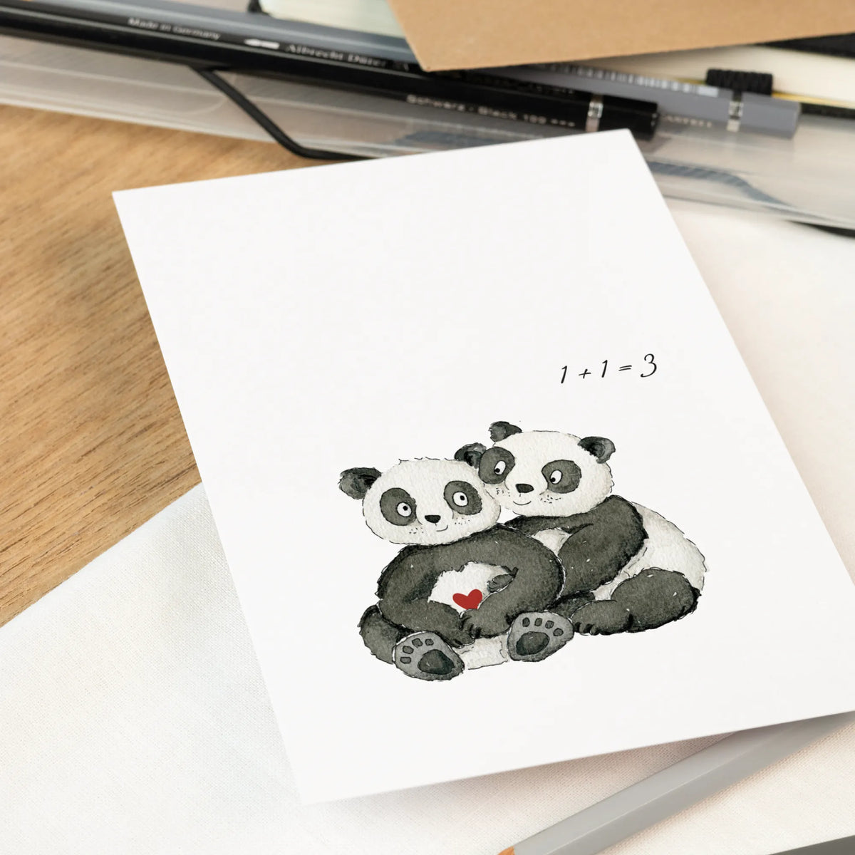 Nahaufnahme der Schwangerschaftskarte mit zwei sich umarmenden handgemalten Pandas, rotem Herz und Schriftzug 1+1=3