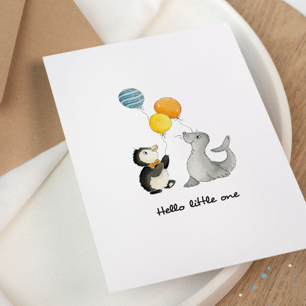 Nahaufnahme der Glückwunschkarte zur Geburt Baby: handgemaltes Aquarell Motiv Pinguin und Seehund mit Luftballons, „Hello little one“ auf weißem Papier.