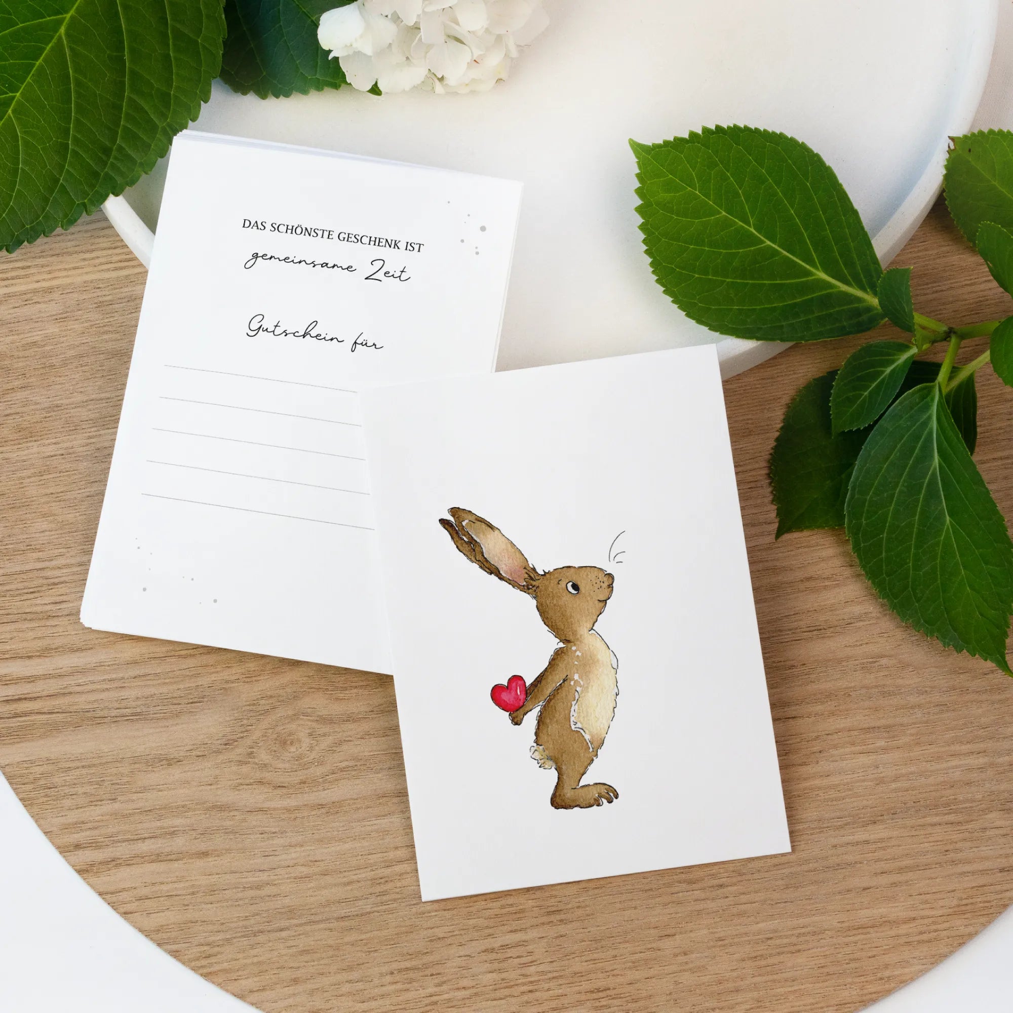 Zwei weiße Gutscheinkarten auf einem Holztisch. Vorne ein handgemaltes Hase Aquarell Motiv und hinten Linien zum Ausfüllen.