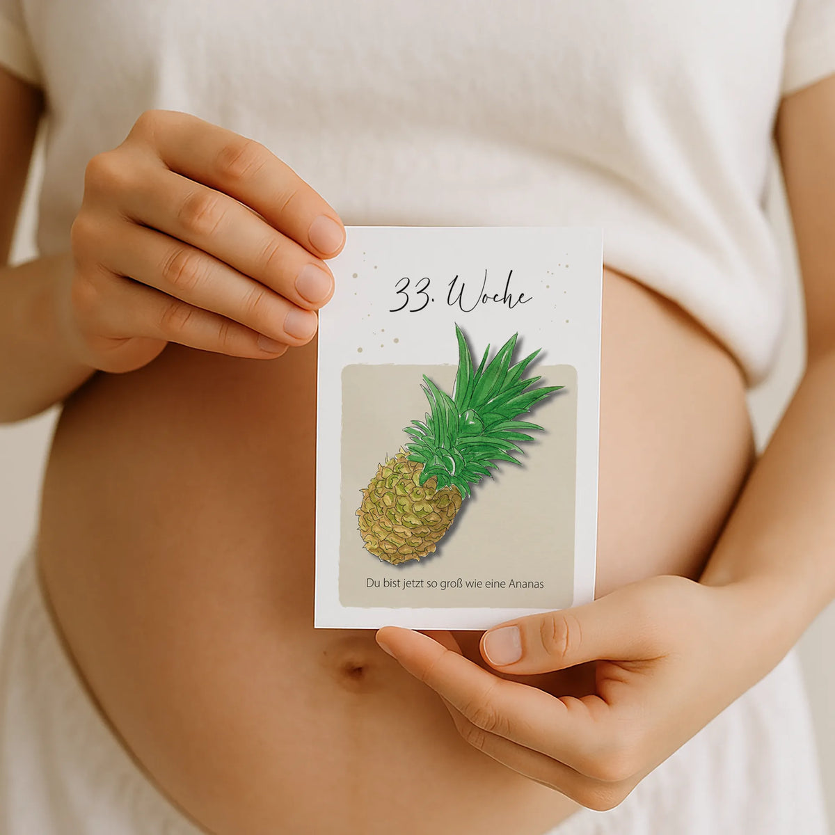 Schwangere hält Meilensteinkarte 33. Woche mit Aquarell Motiv Ananas vor ihren Babybauch