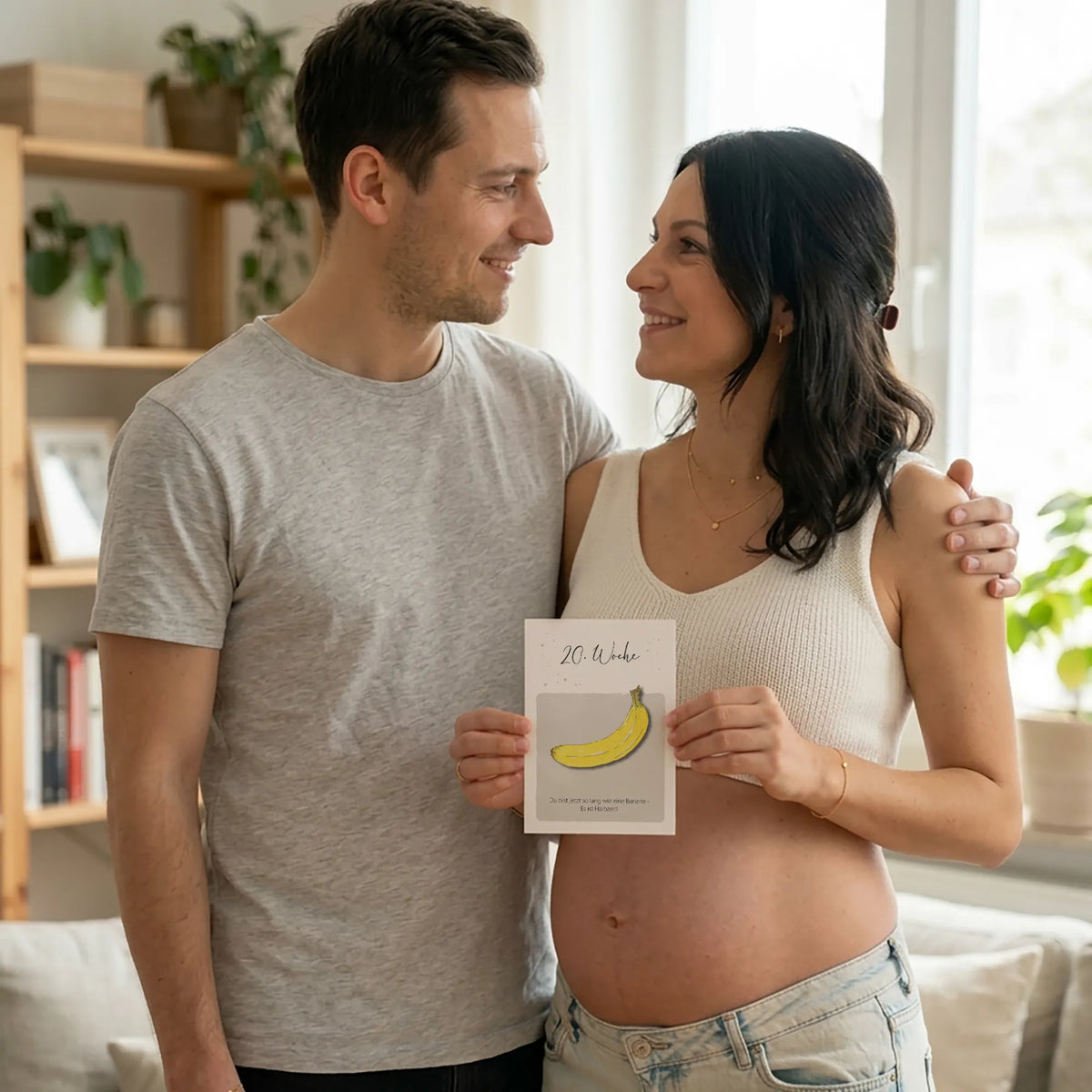 Pärchen mit Karte 20. SSW. Frau hält Karte vor ihren Babybauch, beide schauen sich lächelnd an.