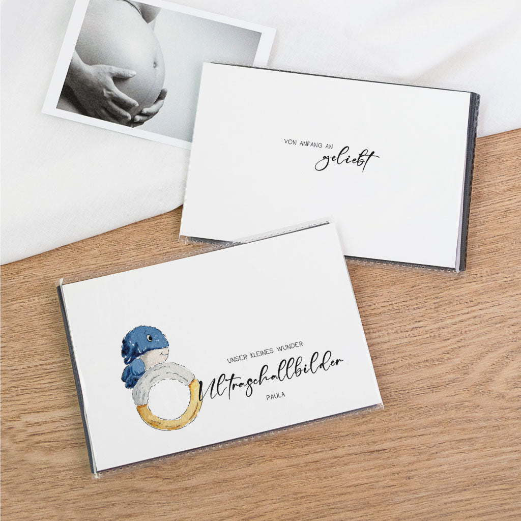 Zwei schwarze Einsteckalben für Ultraschallbilder auf Holzuntergrund – Vorderseite mit Wal-Illustration, Rückseite mit Schriftzug „von Anfang an geliebt“, Ultraschallfoto daneben