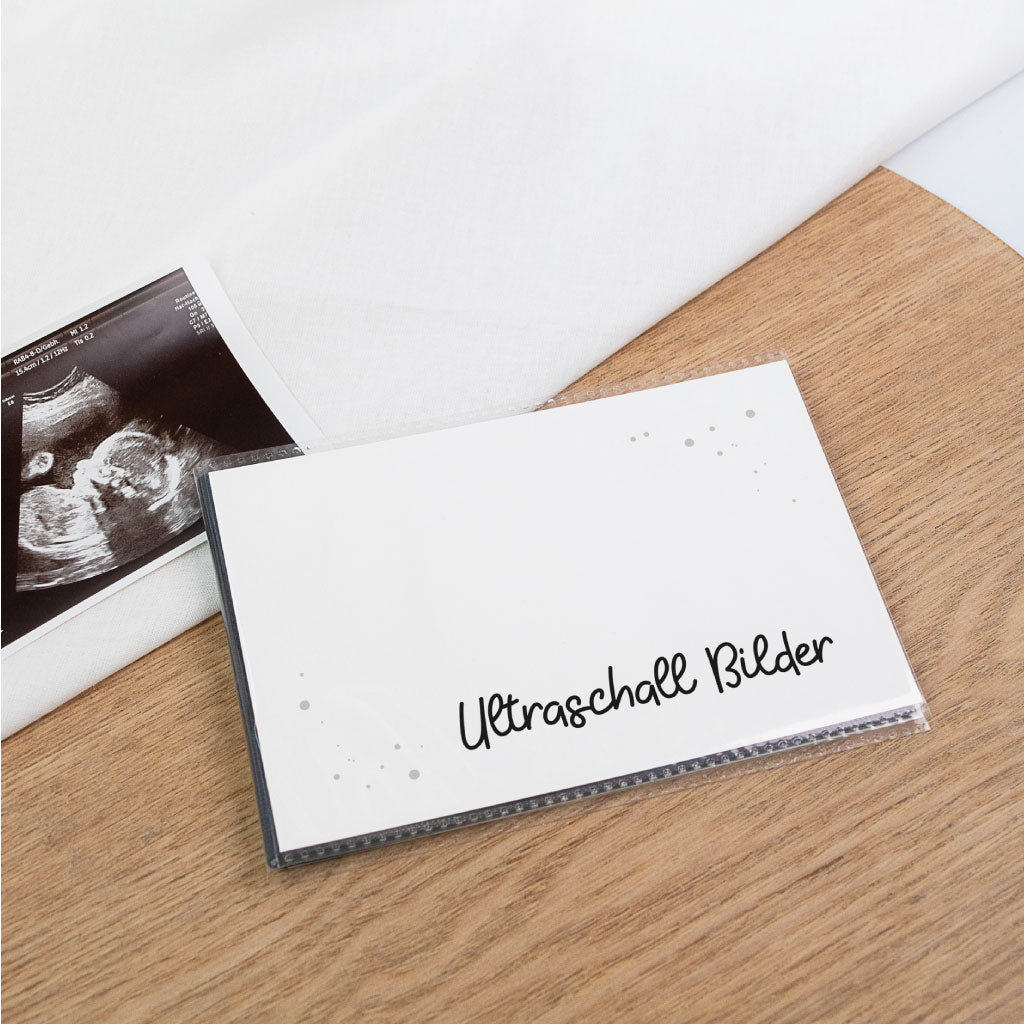 Schwarzes Einsteckalbum zum Schutz für Ultraschall Fotos: weißes Cover mit „Ultraschall Bilder“ auf schwarzem Albumrücken, Ultraschallfoto liegt links daneben.