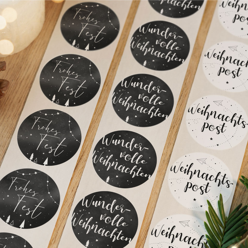 Drei verschiedene Weihnachtsaufkleber Designs auf Rollen nebeneinander, im Hintergrund Tannenzweige und Lichterketten.