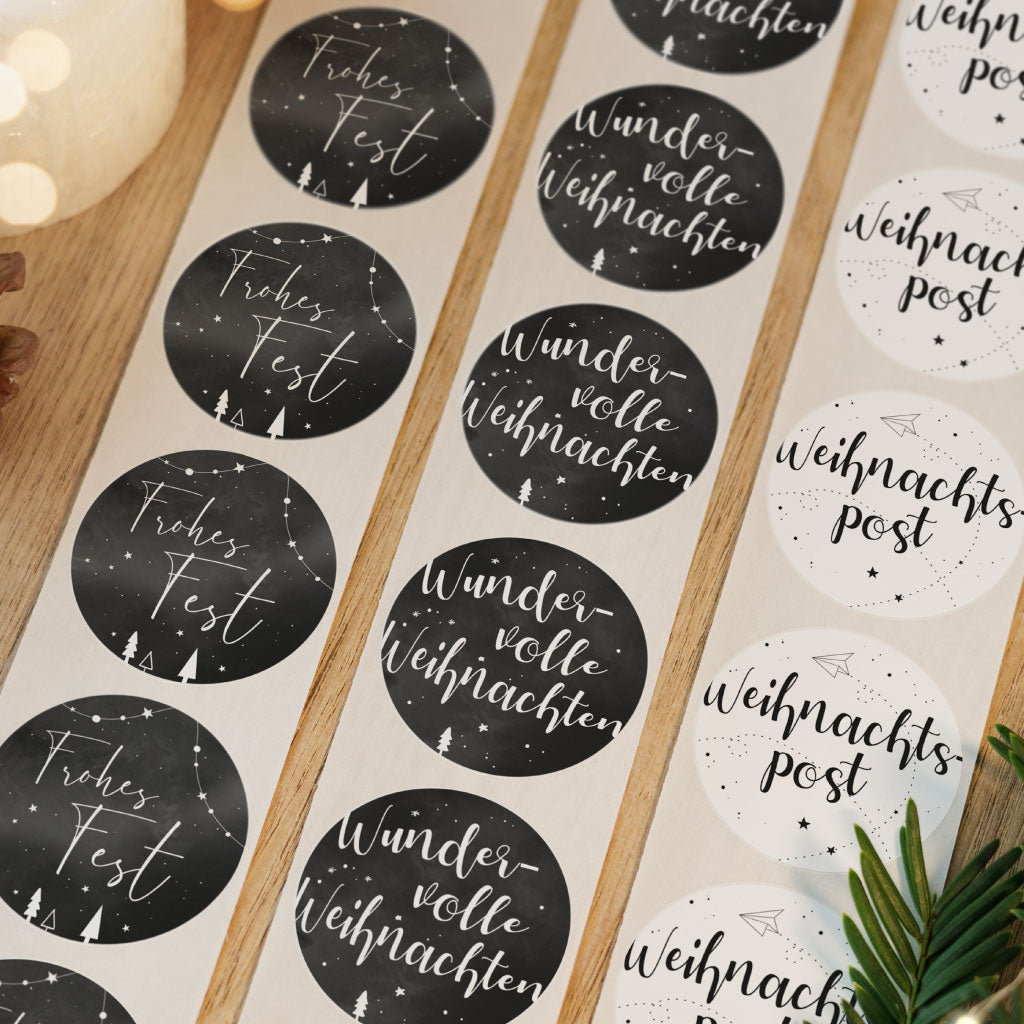 Drei verschiedene Weihnachtsaufkleber Designs auf Rollen nebeneinander, im Hintergrund Tannenzweige und Lichterketten.