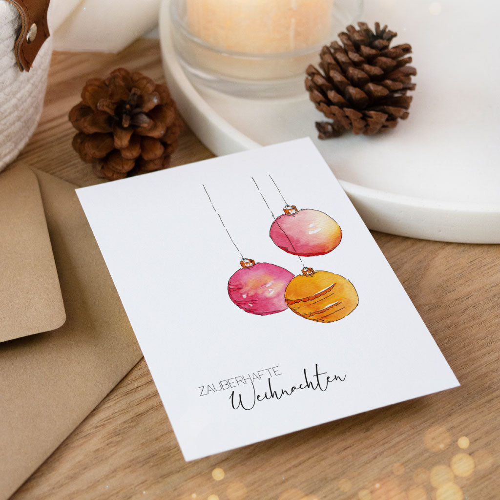 Liebevoll gestaltete Aquarellkarte mit drei weihnachtlichen Christbaumkugeln und dem Gruß „Zauberhafte Weihnachten“, dekorativ arrangiert mit Umschlag und Tannenzapfen.