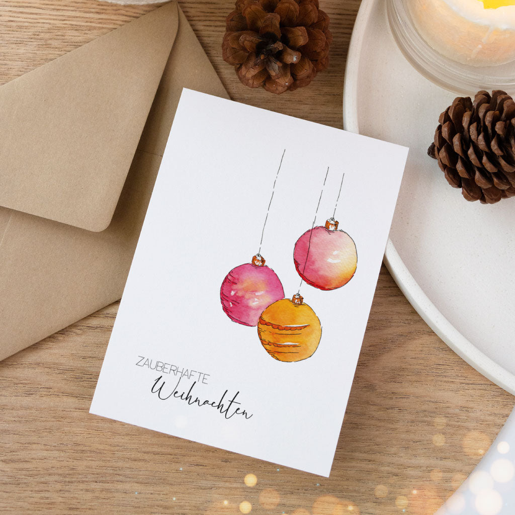 Besondere Postkarte im Aquarell-Stil, zeigt drei Christbaumkugeln in Pink, Orange und Gold sowie den Schriftzug „Zauberhafte Weihnachten“, auf Holzoberfläche präsentiert.