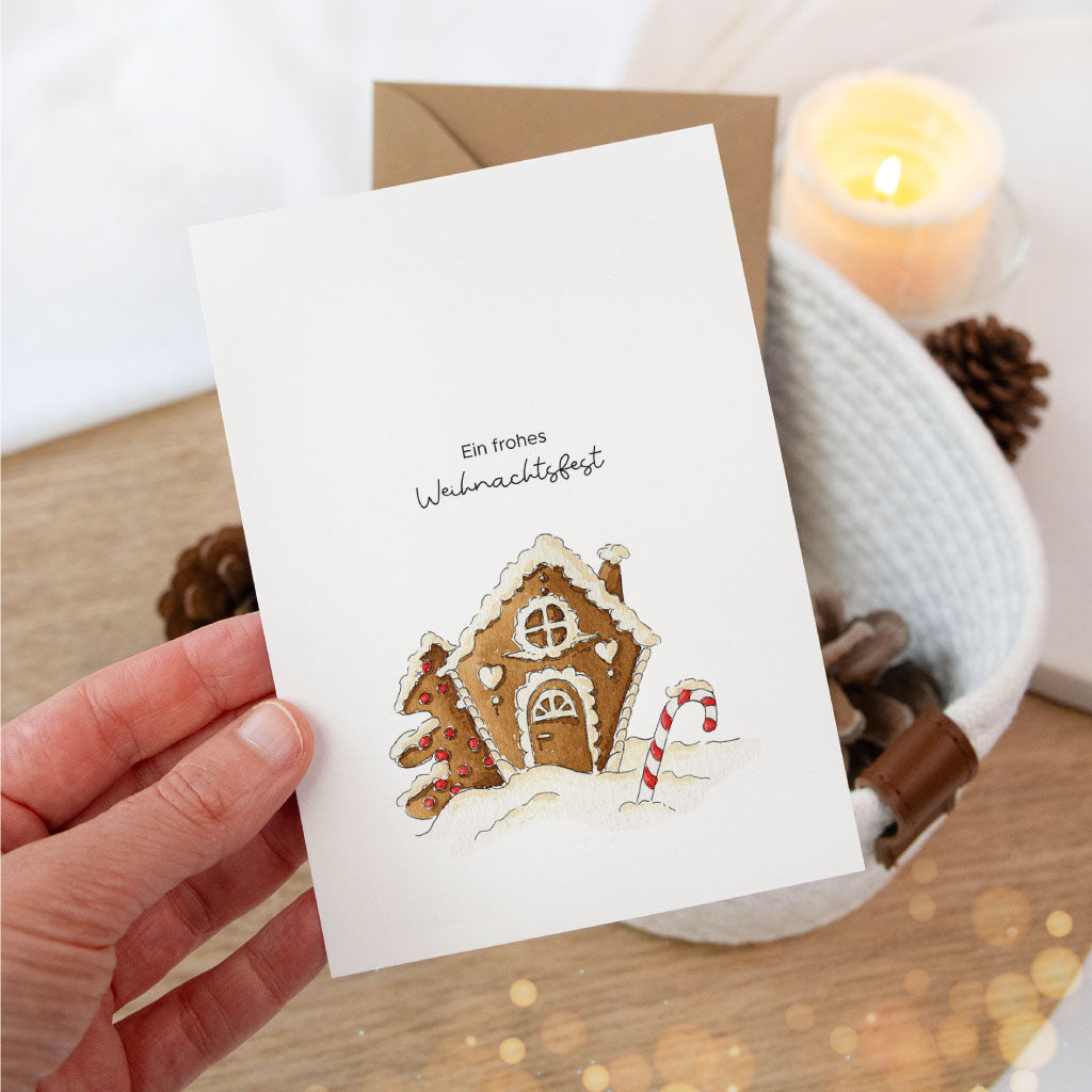 Nahaufnahme einer Hand, die eine Aquarell-Weihnachtskarte mit Lebkuchenhaus-Motiv hält – ideal als Postkarte oder für liebevolle Geschenkverpackungen an Weihnachten.