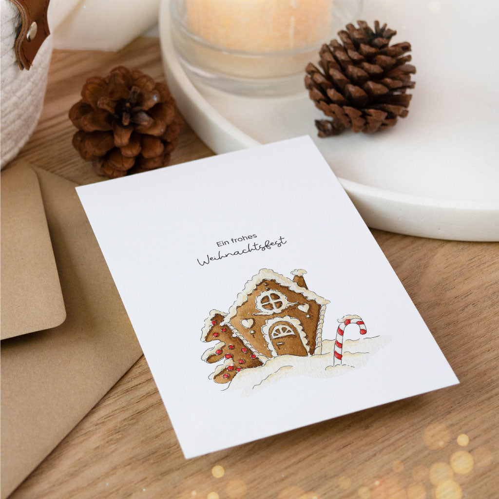 Weihnachtlich inszenierte Postkarte mit Aquarellmotiv eines verschneiten Lebkuchenhauses, daneben Deko mit Kerze und Tannenzapfen – perfekte Ergänzung zu Weihnachtsgeschenken.