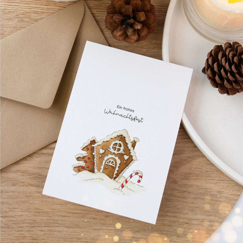 Flach auf Holz liegende Weihnachtskarte mit handgemaltem Aquarell-Lebkuchenhaus, kombiniert mit Umschlag und natürlicher Dekoration.