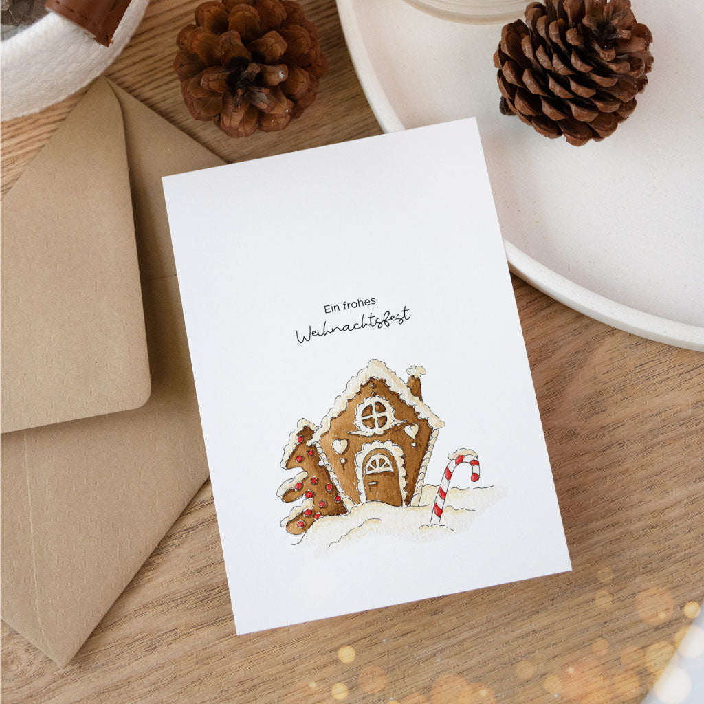 Postkarte mit liebevollem Aquarellmotiv eines Lebkuchenhauses, winterlich dekoriert mit Kerze, Zapfen und Umschlag – ideal für die festliche Geschenkverpackung.