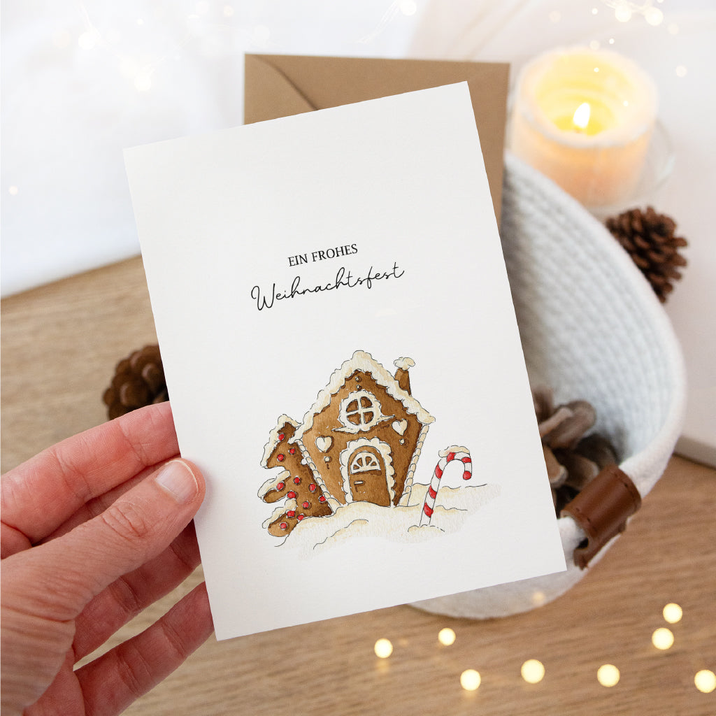 Hand hält eine Weihnachtskarte mit Lebkuchenhaus Aquarell Motiv vor einem festlich dekorierten Tisch.