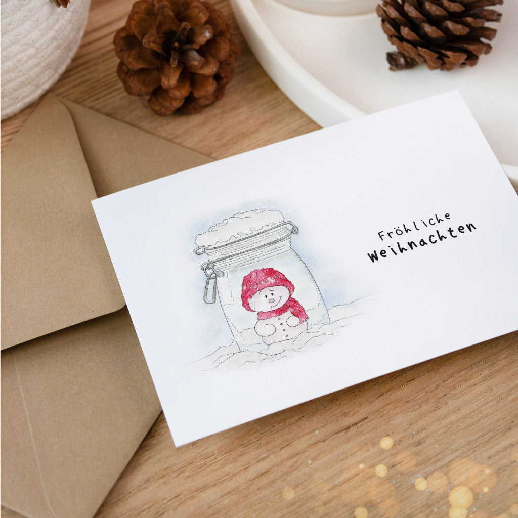 Aquarell-Weihnachtskarte mit niedlichem Schneemann im Glas und handgeschriebenem Schriftzug „Fröhliche Weihnachten“, ideale Ergänzung zu Weihnachtsgeschenken.