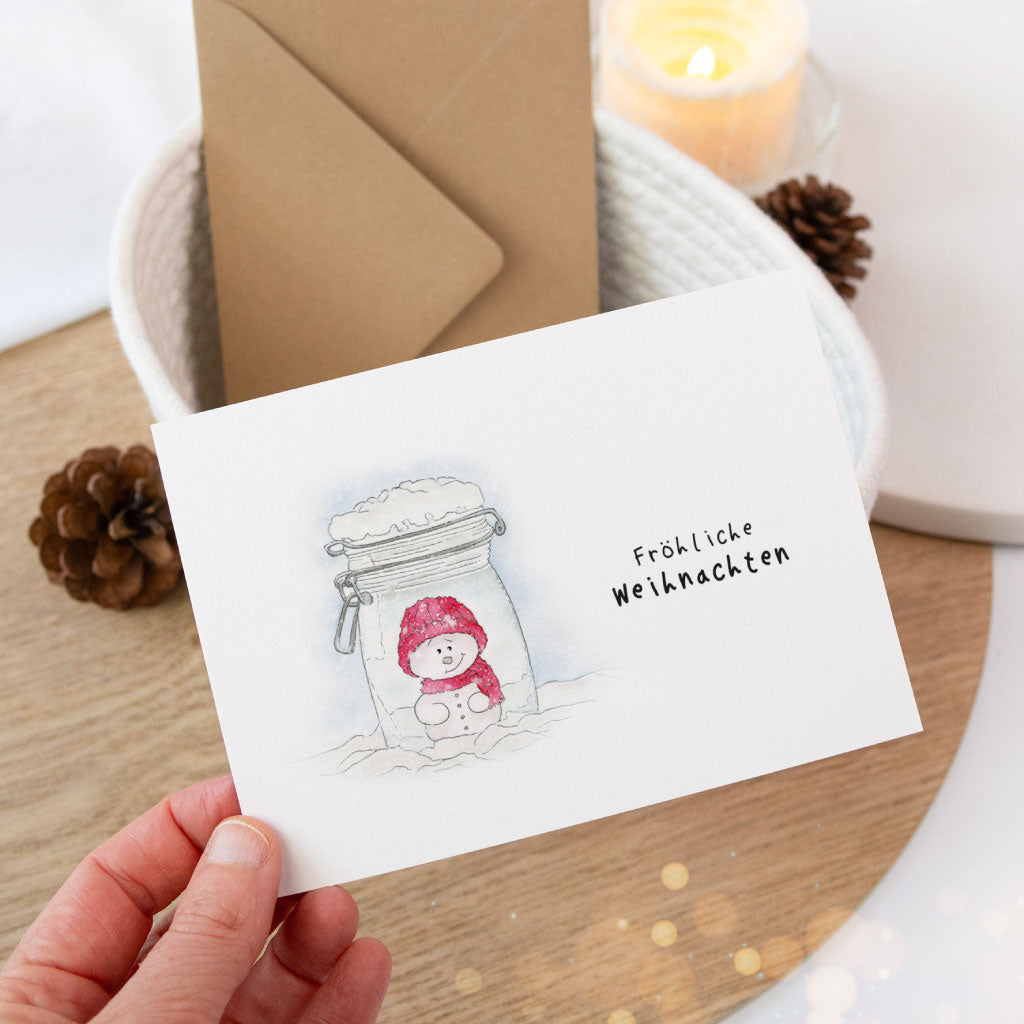 Weihnachtskarte mit Schneemann-Aquarell in stimmungsvoller Deko mit Zapfen und Kerze, handgefertigt und liebevoll illustriert.
