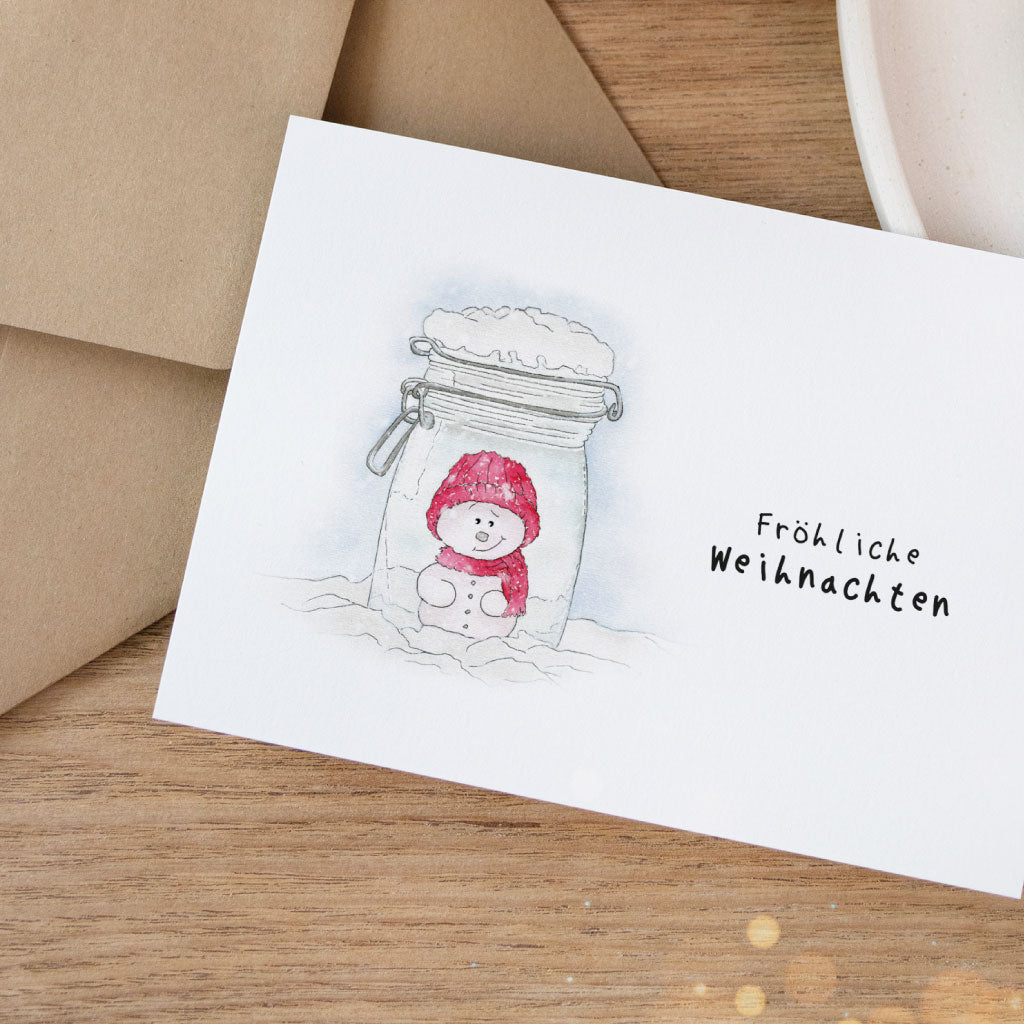 Fröhliche Weihnachtskarte mit süßem Schneemann im Glas, festlich inszeniert – ideale Karte zum Verschenken oder Beilegen zu Geschenken.