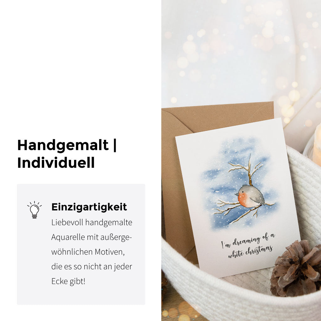Handgemalte Weihnachtskarte mit winterlicher Szene und Rotkehlchen, präsentiert im Korb neben Kerze und Zapfen – einzigartiges Aquarellmotiv für festliche Grüße.