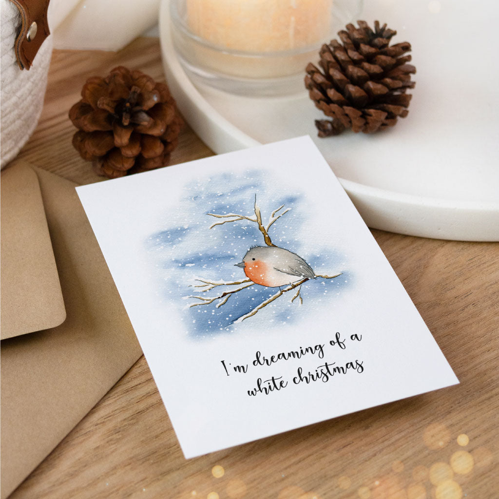 Detailaufnahme der Weihnachtskarte mit handgemaltem Aquarell eines Rotkehlchens, liegend auf einem Holzuntergrund, umgeben von weihnachtlicher Dekoration.