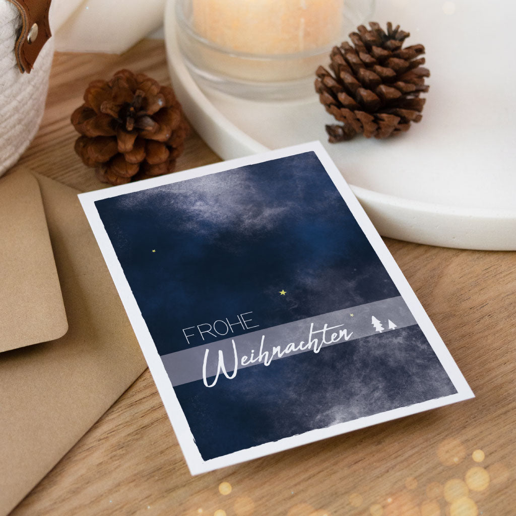 Aquarell-Weihnachtskarte mit minimalistischer Gestaltung und weihnachtlichem Nachthimmel, dekorativ auf Holzuntergrund mit Umschlag und Naturdeko.