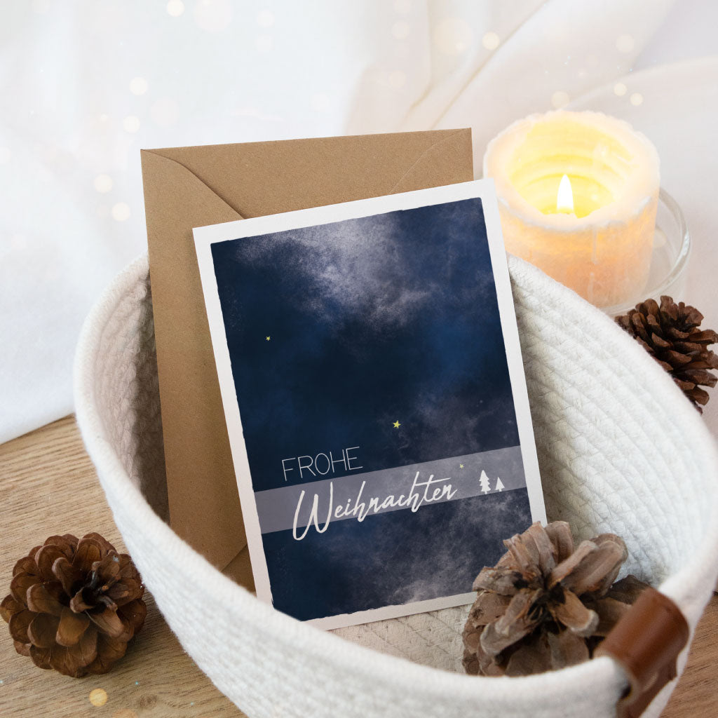 Handgemalte Klappkarte mit dunkelblauem Aquarellhimmel und feinen Schneeflocken, liebevoll im Stoffkorb mit Kerzenlicht und Tannenzapfen inszeniert.