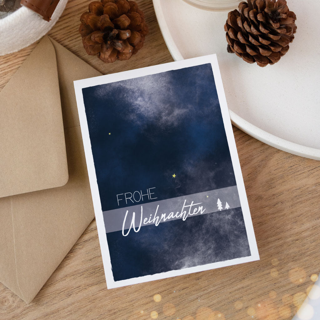 Minimalistische Aquarellkarte mit weihnachtlichem Nachthimmel und dem Text „Frohe Weihnachten“, stilvoll präsentiert auf einem Holztisch mit Naturdeko.