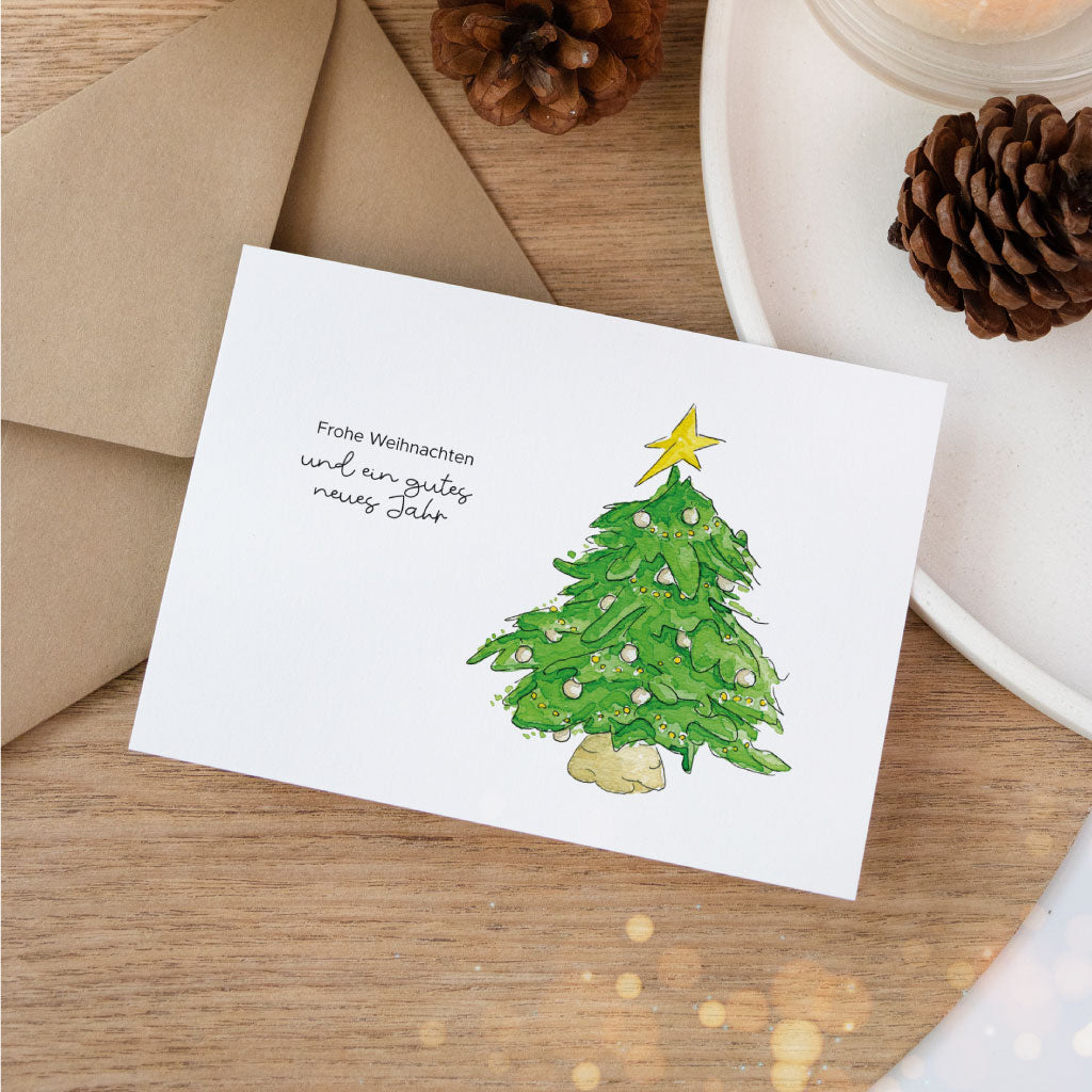 Weihnachtskarte mit handgemaltem Aquarell eines festlich geschmückten Weihnachtsbaums und dem Spruch „Frohe Weihnachten und ein gutes neues Jahr“, liebevoll inszeniert mit Umschlag und Tannenzapfen.