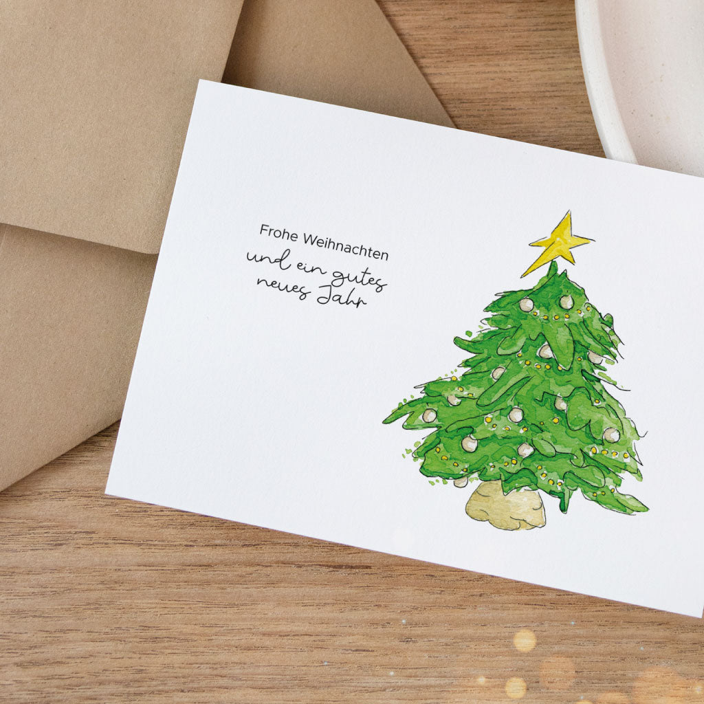 Illustrative Postkarte mit Aquarell-Weihnachtsbaum und dem Spruch „Frohe Weihnachten und ein gutes neues Jahr“, daneben naturfarbener Umschlag auf Holzuntergrund.