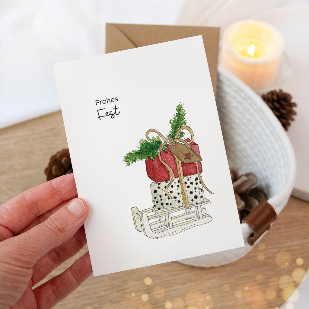 Detailaufnahme der Postkarte „Frohes Fest“ mit Schlitten voller Geschenke, gehalten in der Hand – ideal für persönliche Weihnachtsgrüße oder zum Geschenke verpacken.
