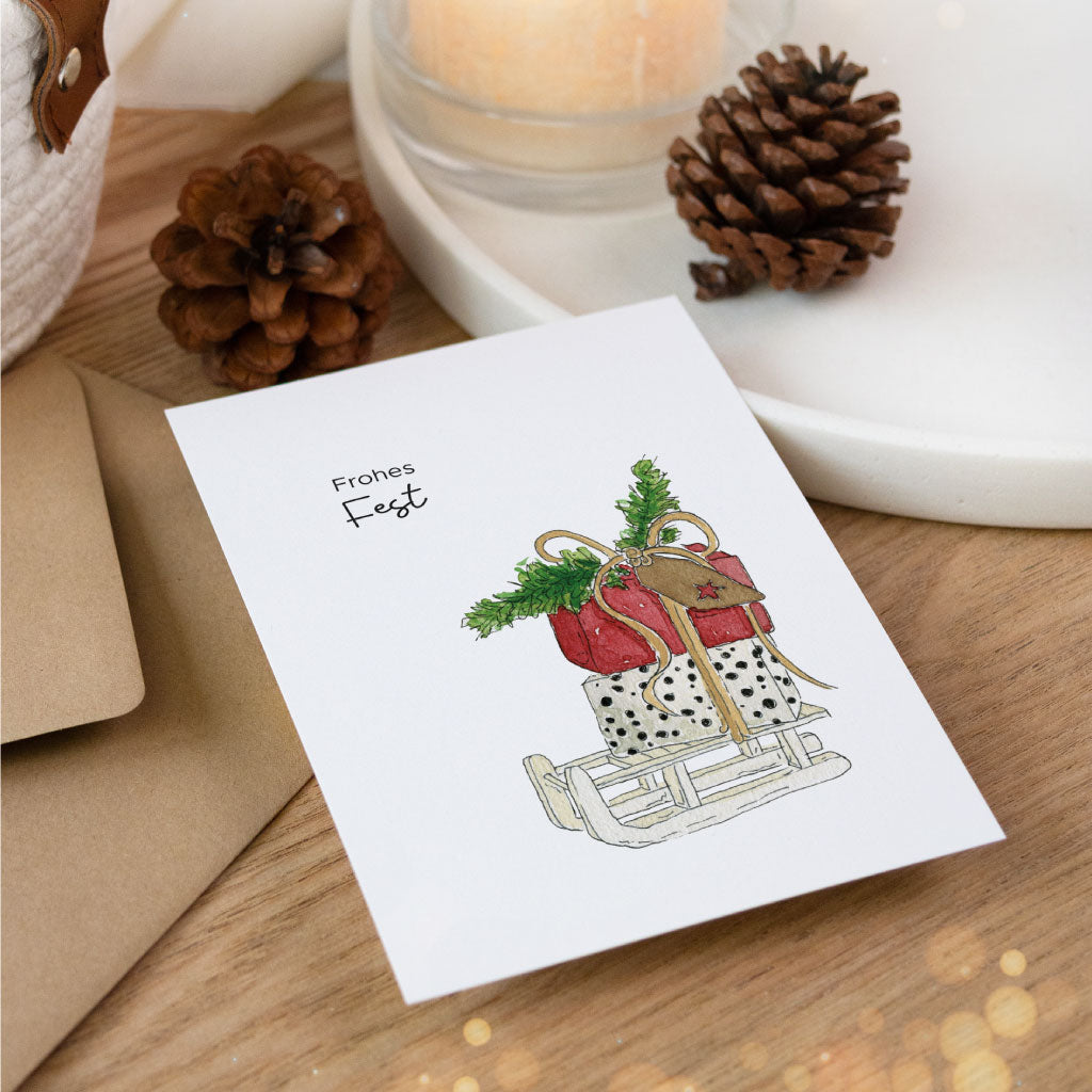 Stimmungsvoll arrangierte Aquarell-Weihnachtskarte mit rotem Geschenkpaket auf einem Schlitten und dem Spruch „Frohes Fest“, kombiniert mit Kerze und Tannenzapfen.