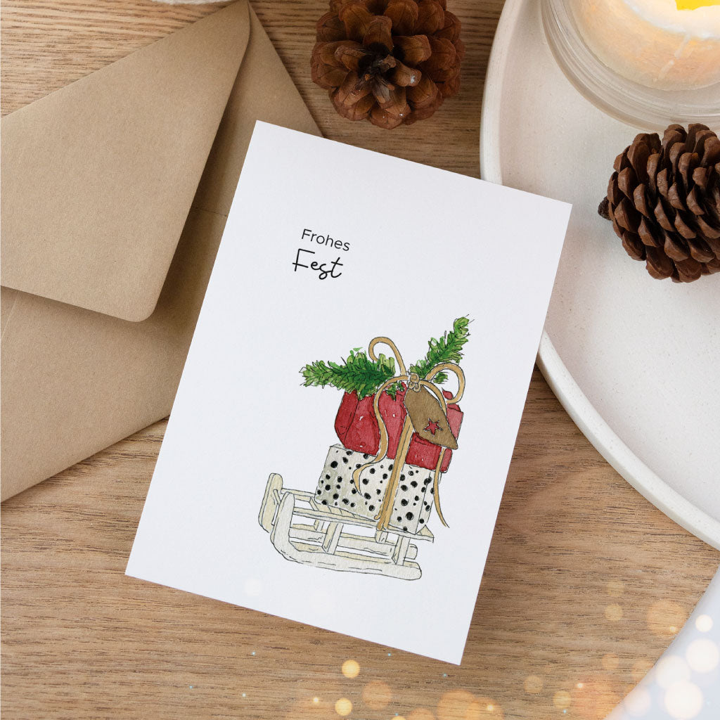 Postkarte mit winterlichem Motiv: Schlitten mit gepackten Geschenken und Zweigen, daneben naturfarbener Umschlag – ideal als liebevolle Weihnachtskarte.