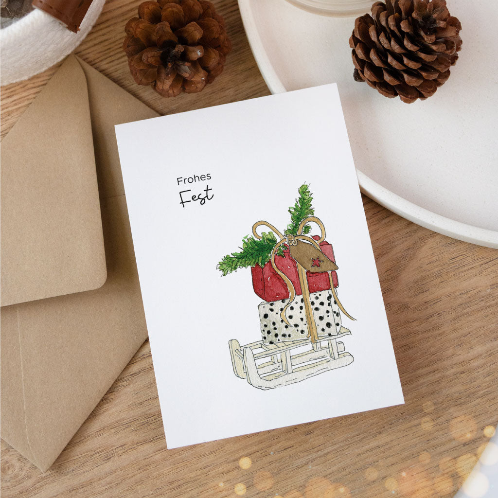 Illustrative Weihnachtskarte mit handgemaltem Aquarell eines Geschenkschlittens und der Aufschrift „Frohes Fest“, dekorativ auf Holzuntergrund platziert.